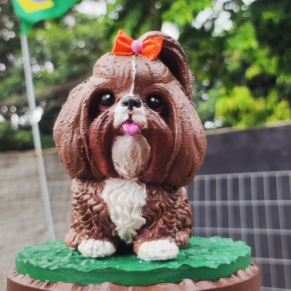 Miniatura de uma shih-tzu marrom e branca com laço laranja, sobre base de grama com o nome CACAU
